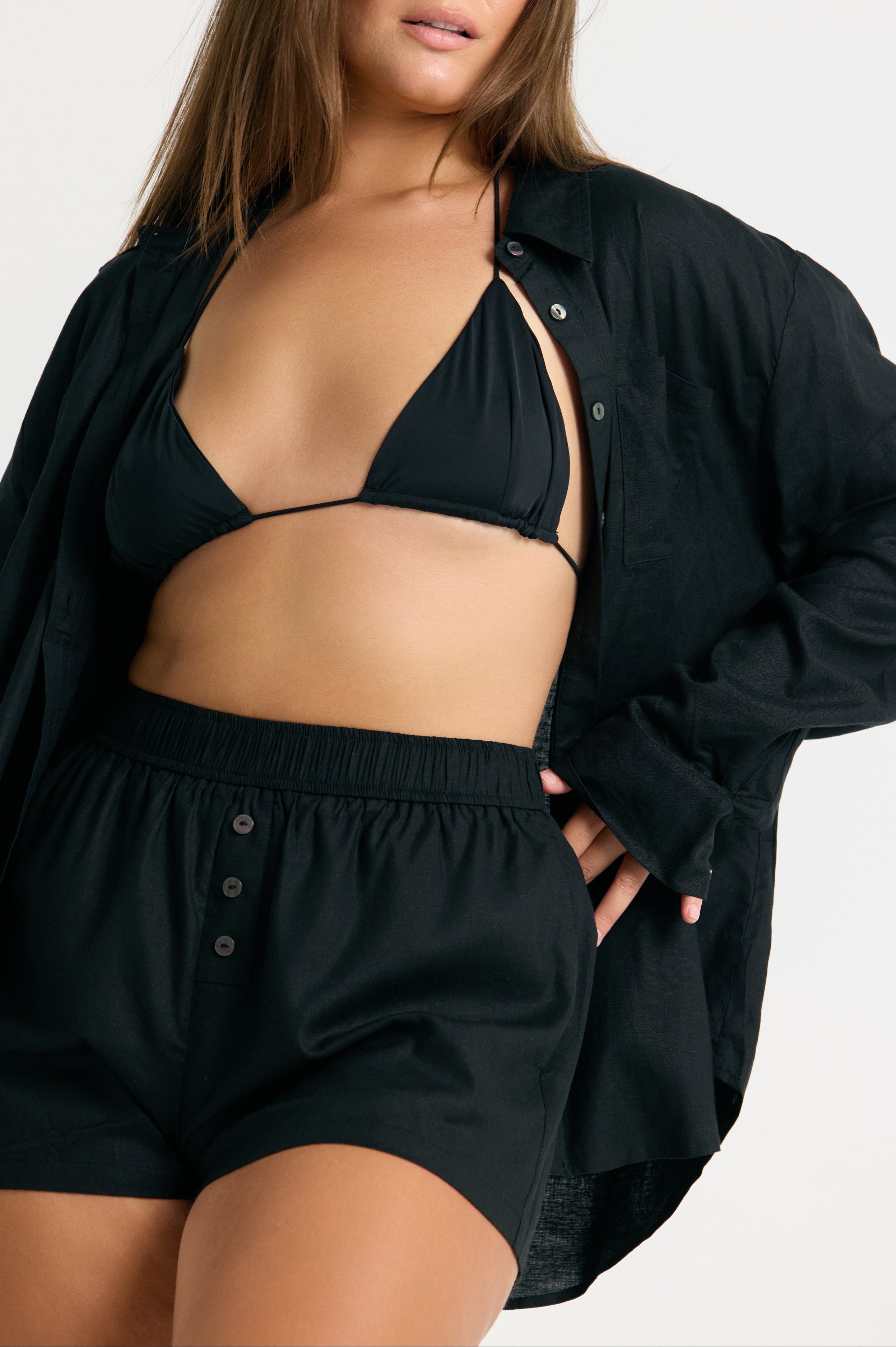 Woman wearing a black linen top and linen shorts with a black bikini top on a white background