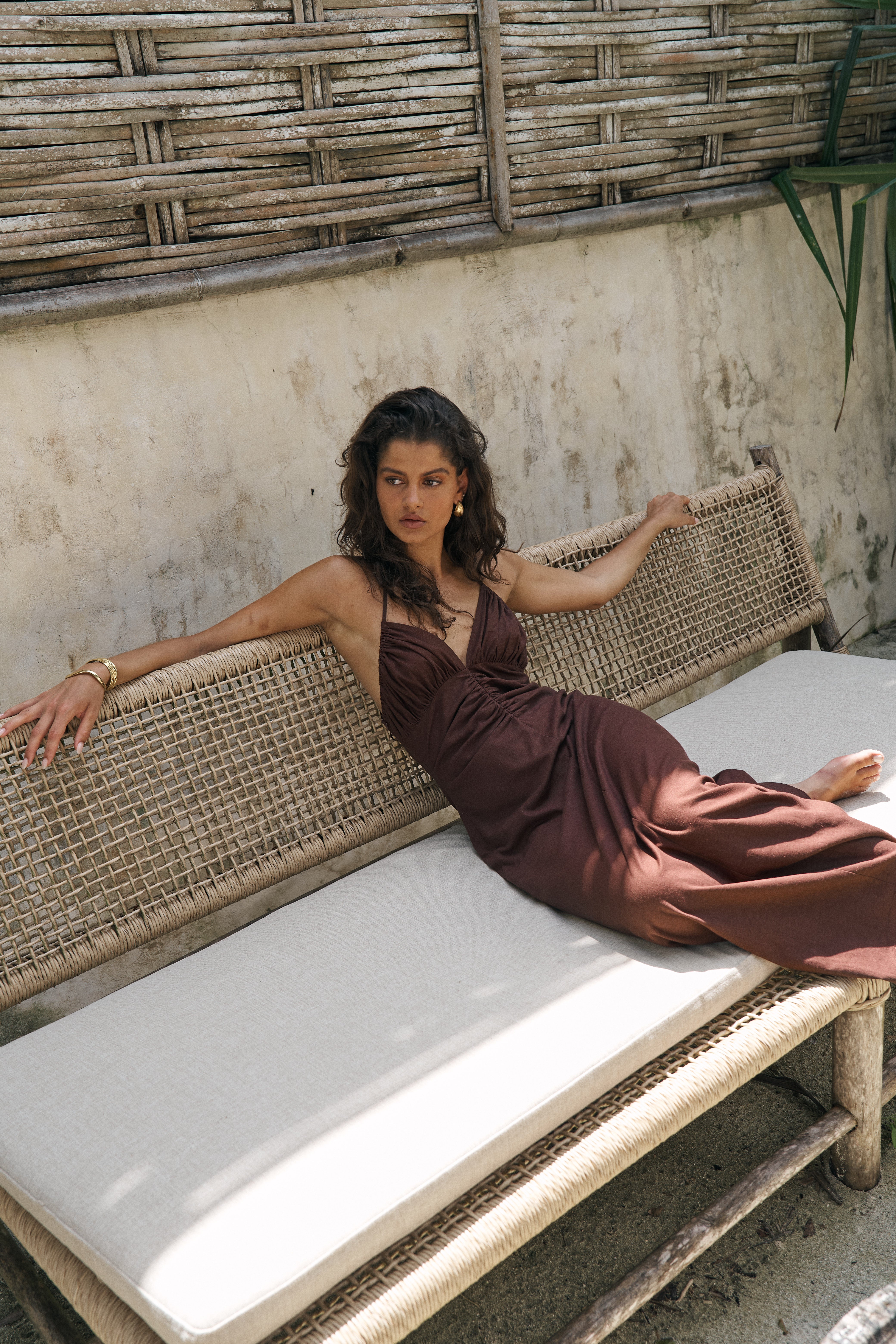 Woman in a brown dress sitting on a wicker chair with a textured wall and plant in the background
