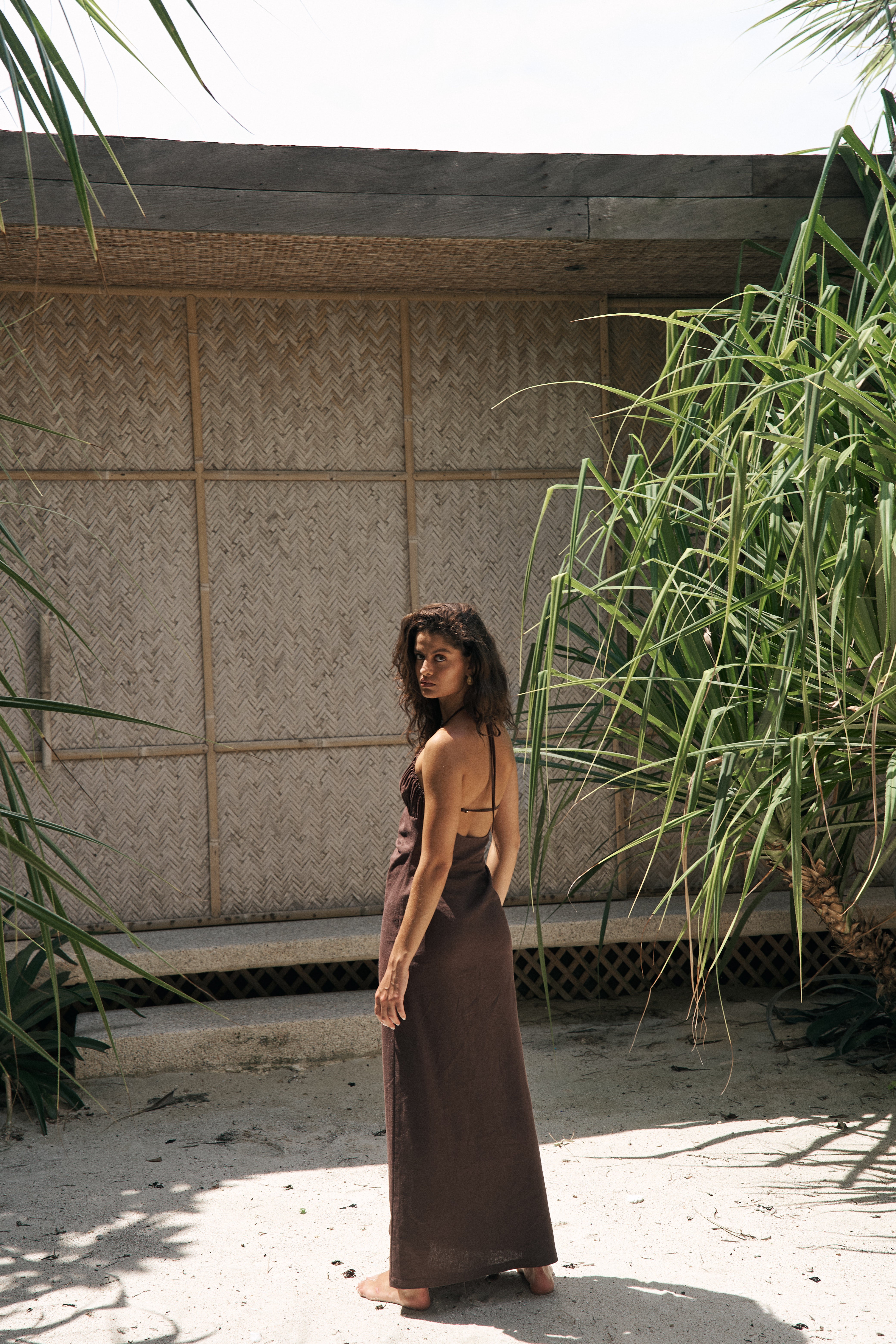 Woman in a long brown dress standing in a natural setting with plants and a wooden structure.
