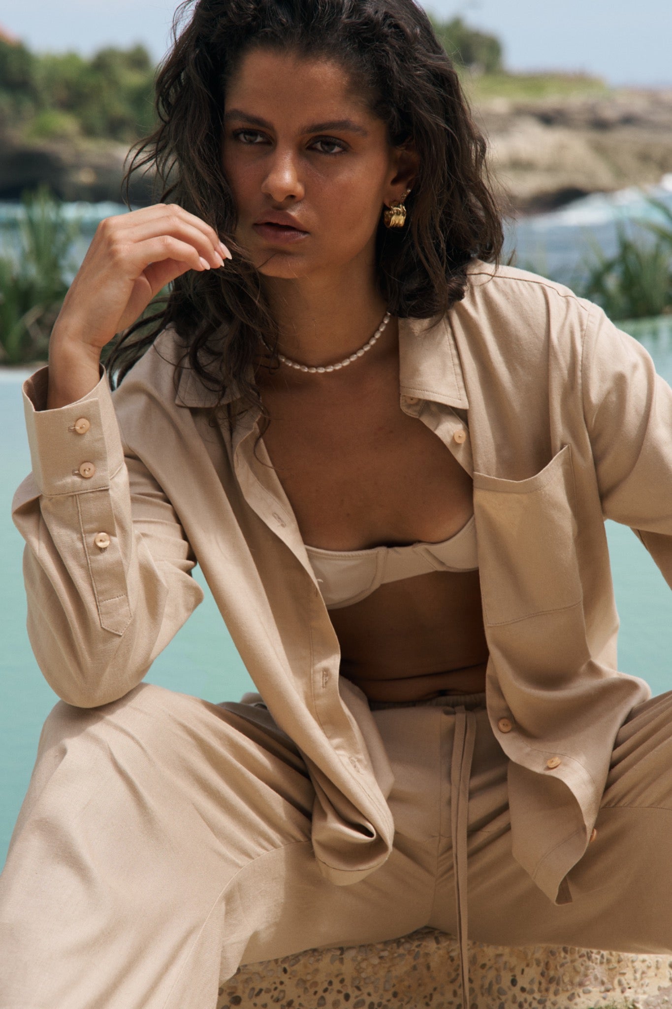 Woman in beige outfit sitting by a body of water with greenery in the background