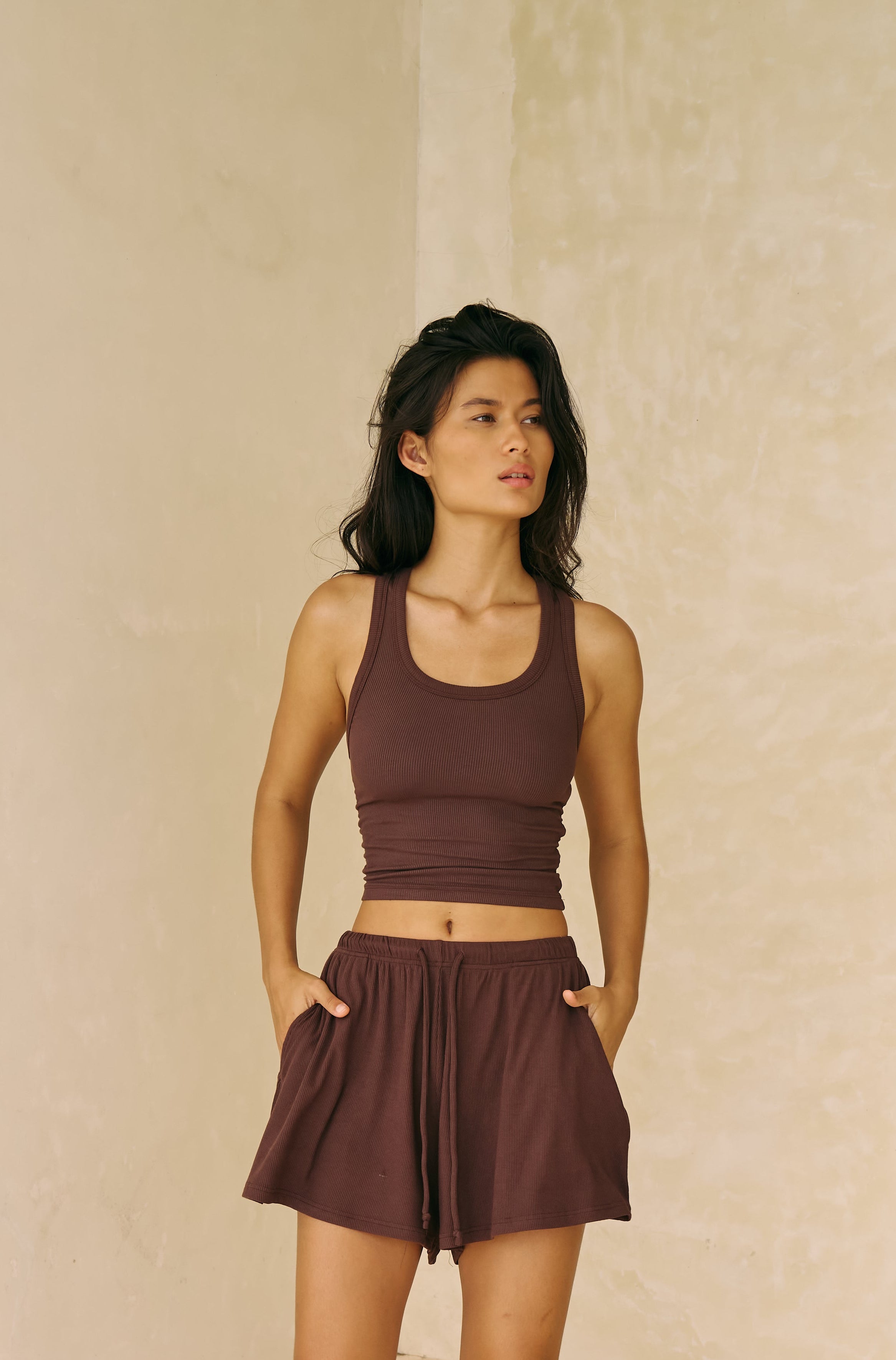 Woman in a brown tank and shorts standing in a sunlit room with columns and steps.