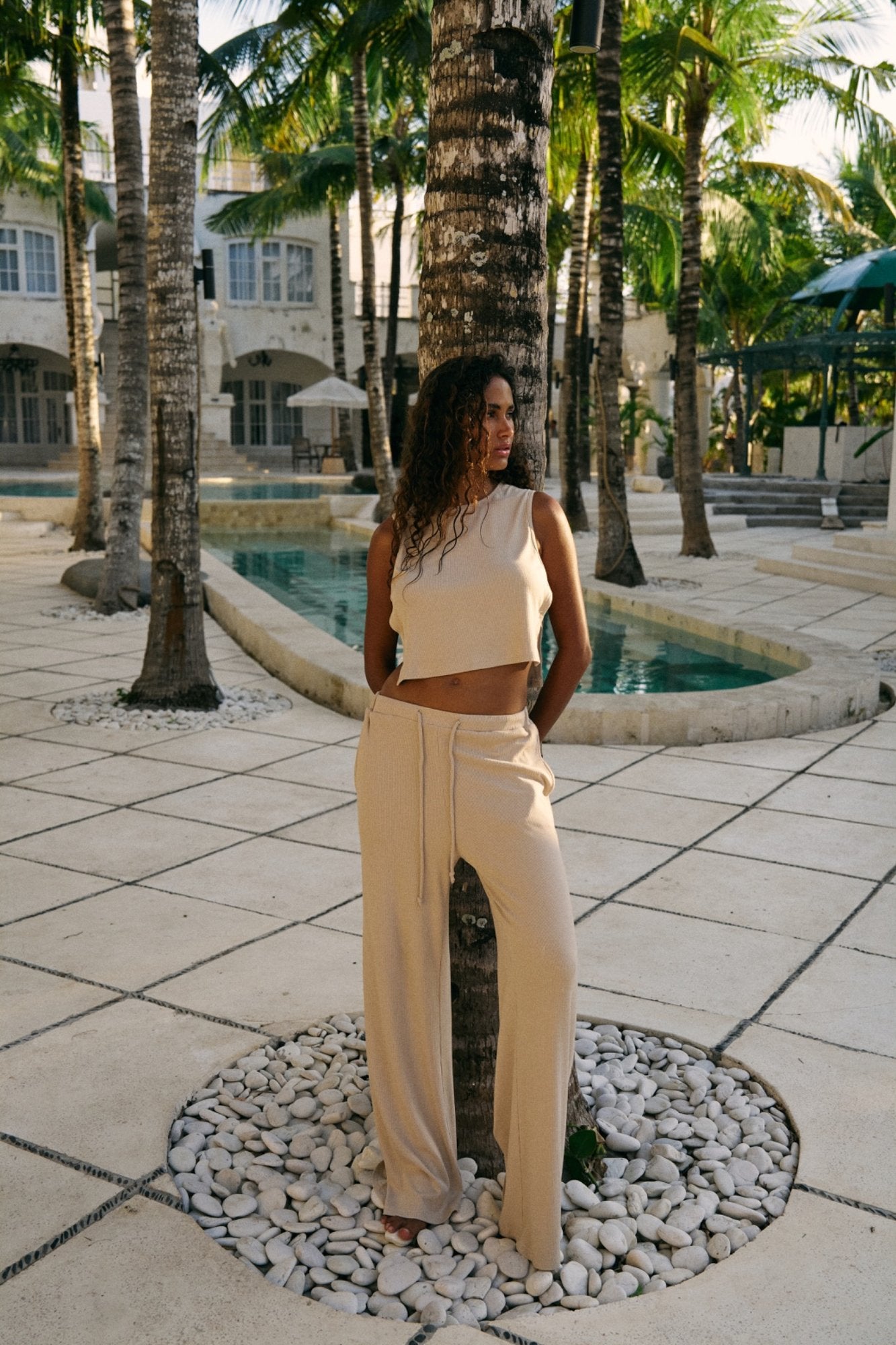 Woman in a coconut/beige top and pants standing in a tropical setting with palm trees and a pool.