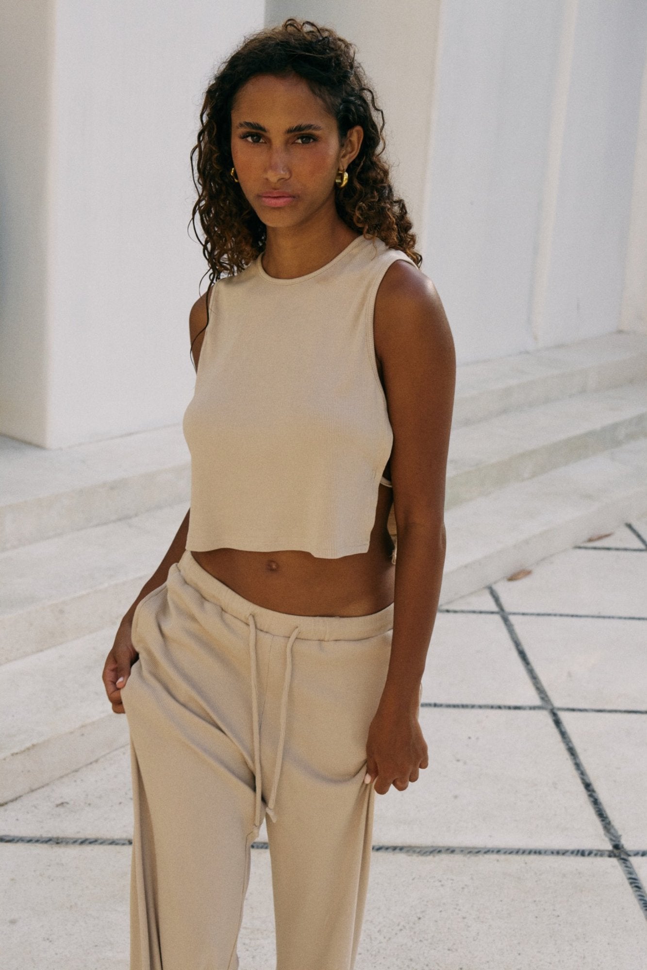 Woman wearing a beige/coconut  sleeveless top and pants standing on a stone pavement.