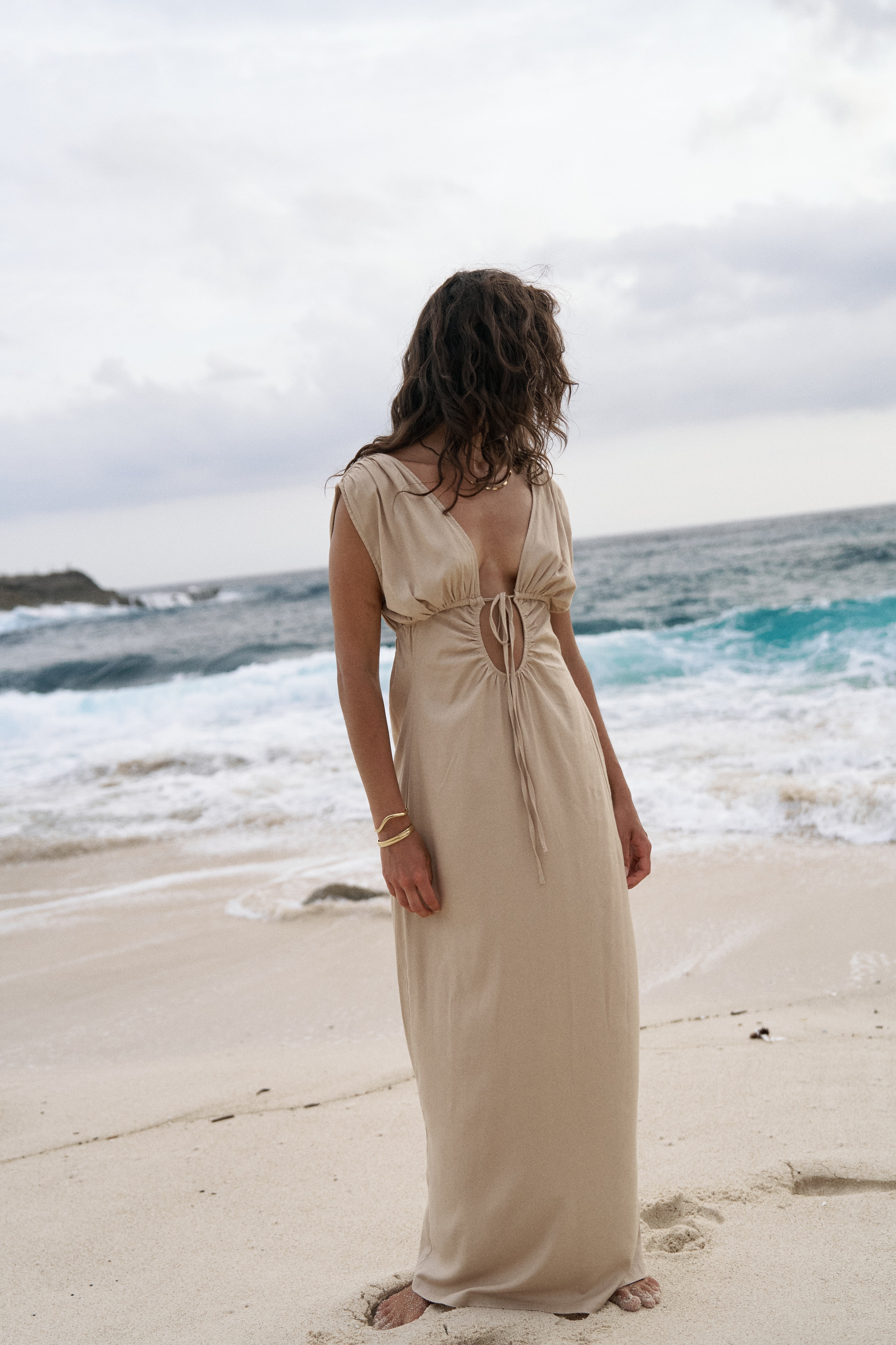 Woman in a beige dress standing on a beach with ocean waves in the background