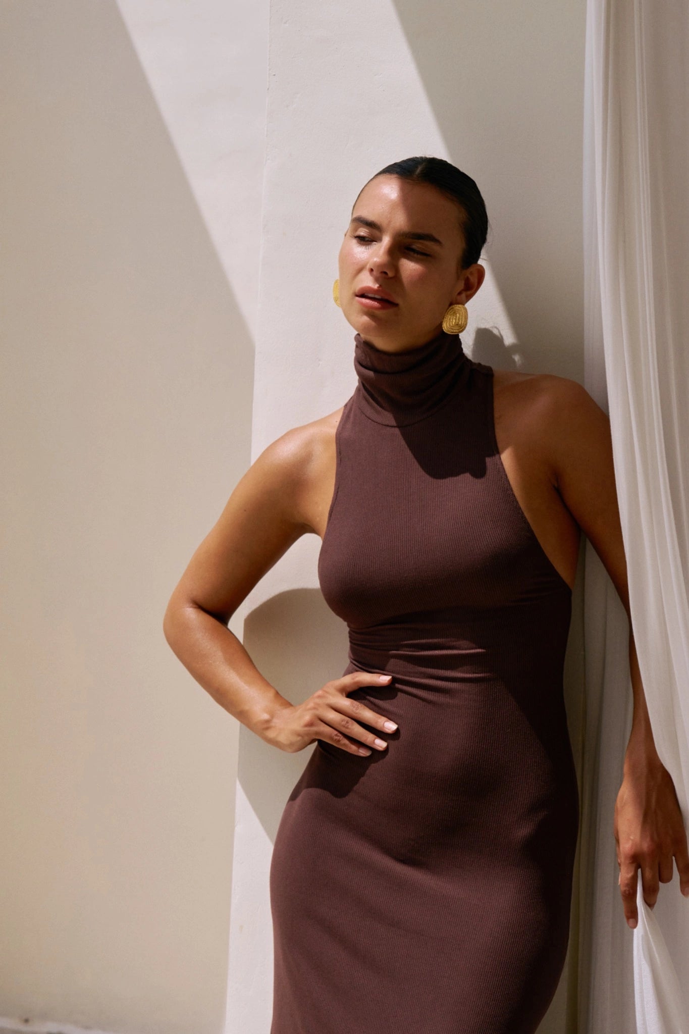 Woman wearing a brown dress standing against a white wall.