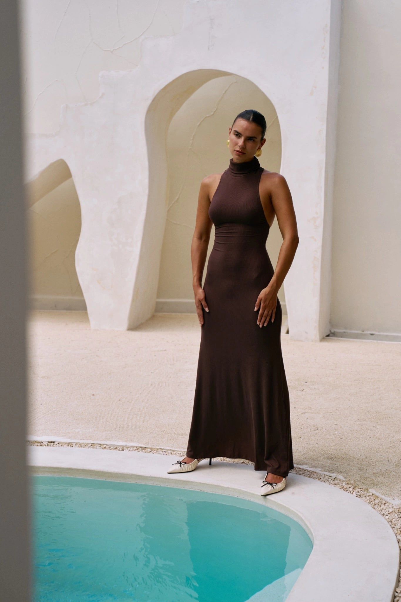 Woman in a brown dress standing by a pool with a white architectural background