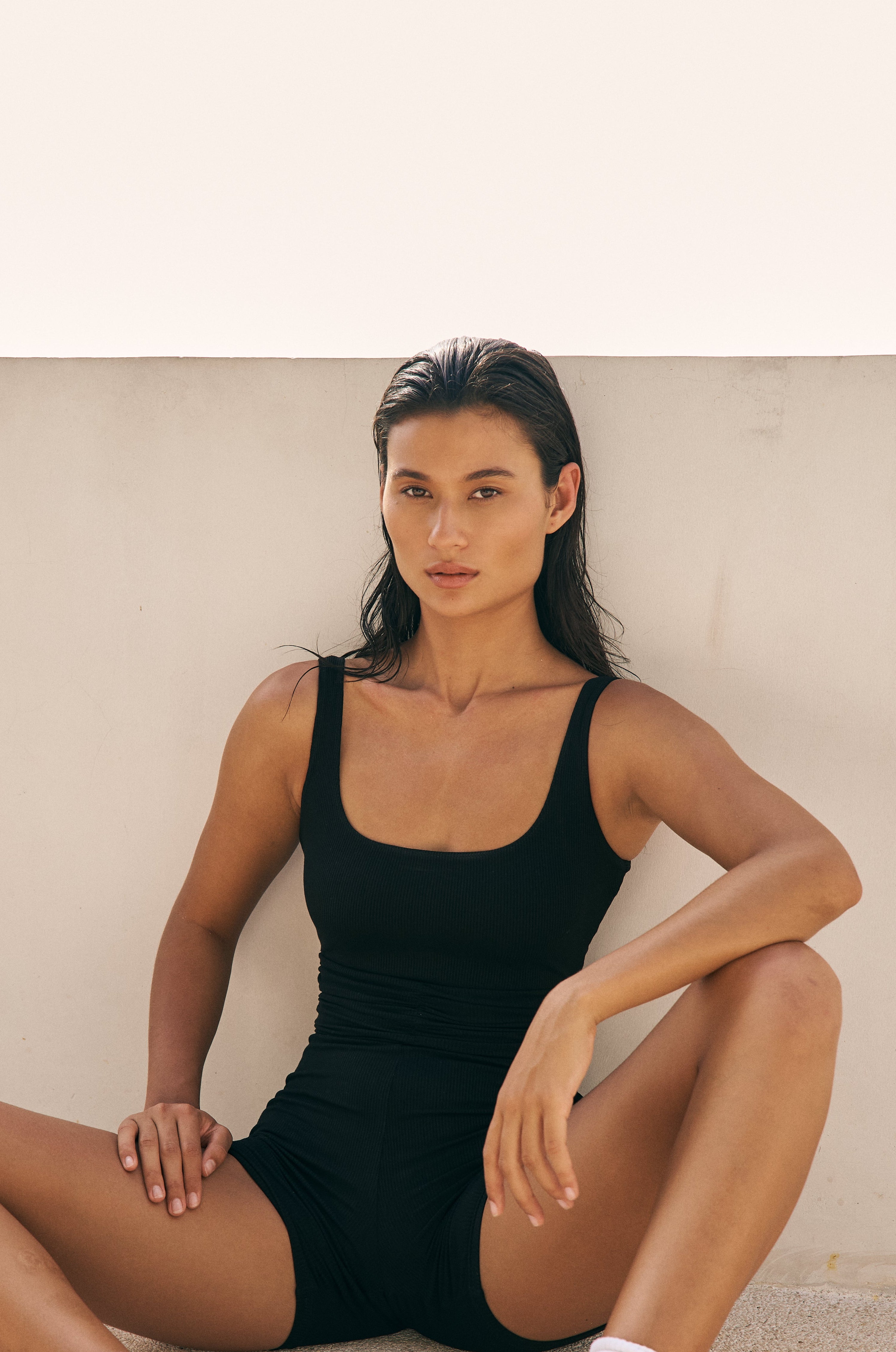 Woman seated on the ground in the Gia black ruched romper, legs stretched out, showing the ribbed texture and gathered waist detail.
