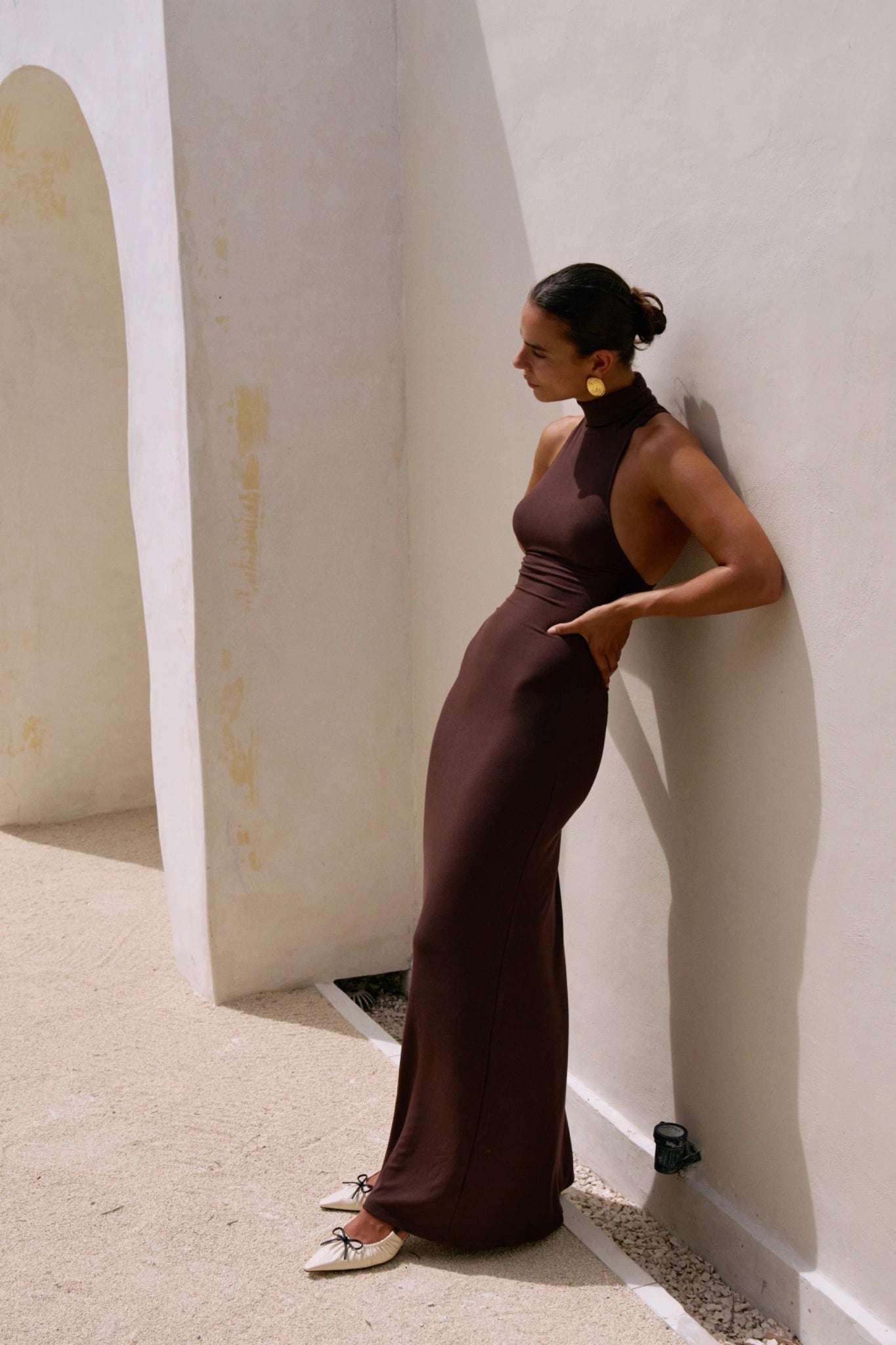Woman in a long brown dress standing against a white wall.