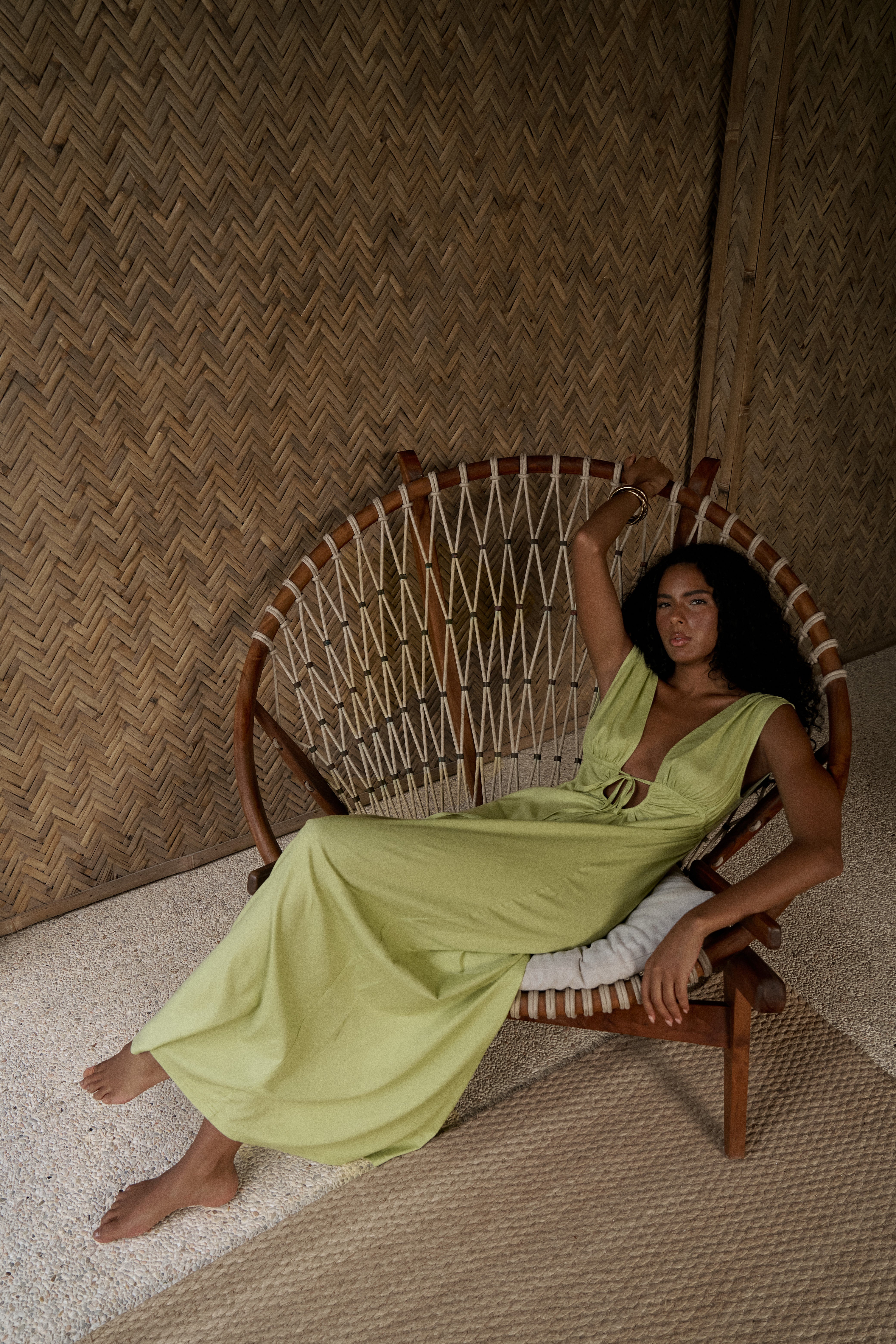 Woman in a green dress sitting on a wooden chair against a woven wall.