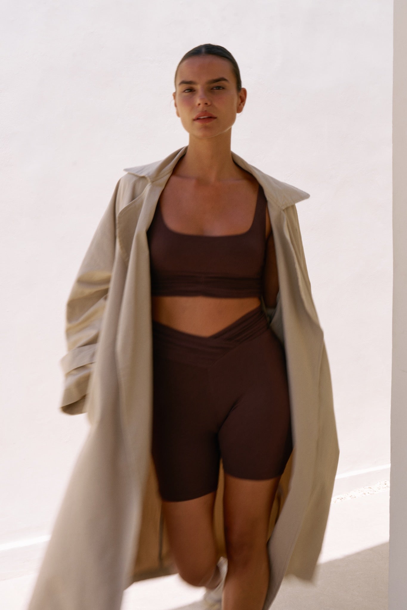 Woman walking outdoors wearing the Demi Top and Demi Bottom in Chocolate, styled with a long beige trench coat