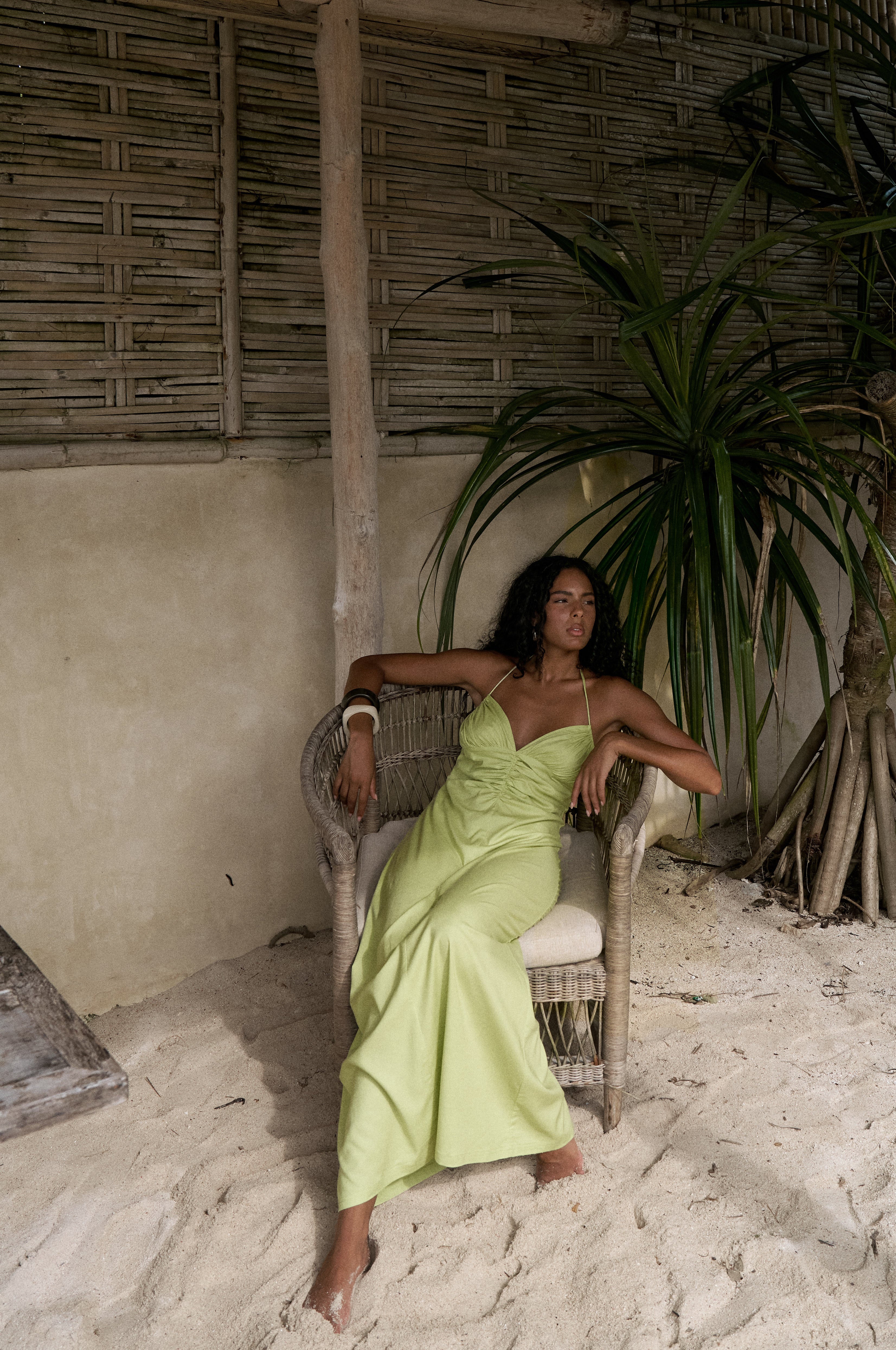 Woman in a green dress sitting in a chair under a rustic wooden structure with plants.