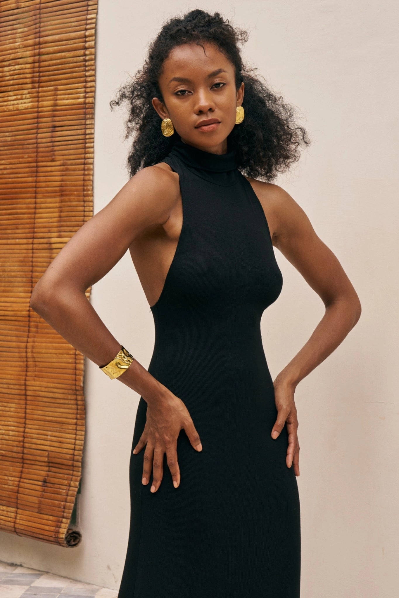 Woman wearing a black dress with gold jewelry against a neutral background