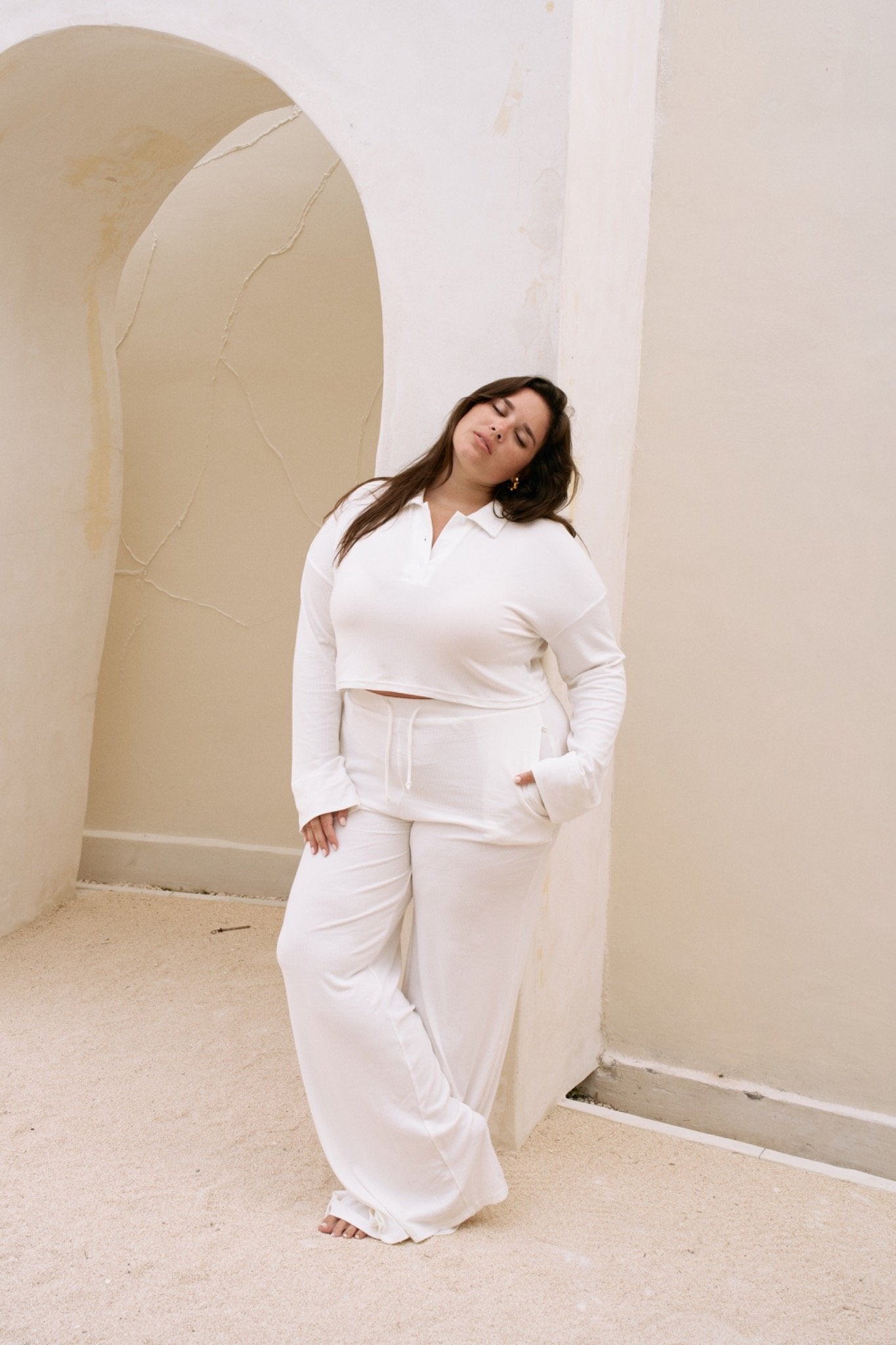Woman in a white  long sleeve top and pants standing in a room with arched doorways.