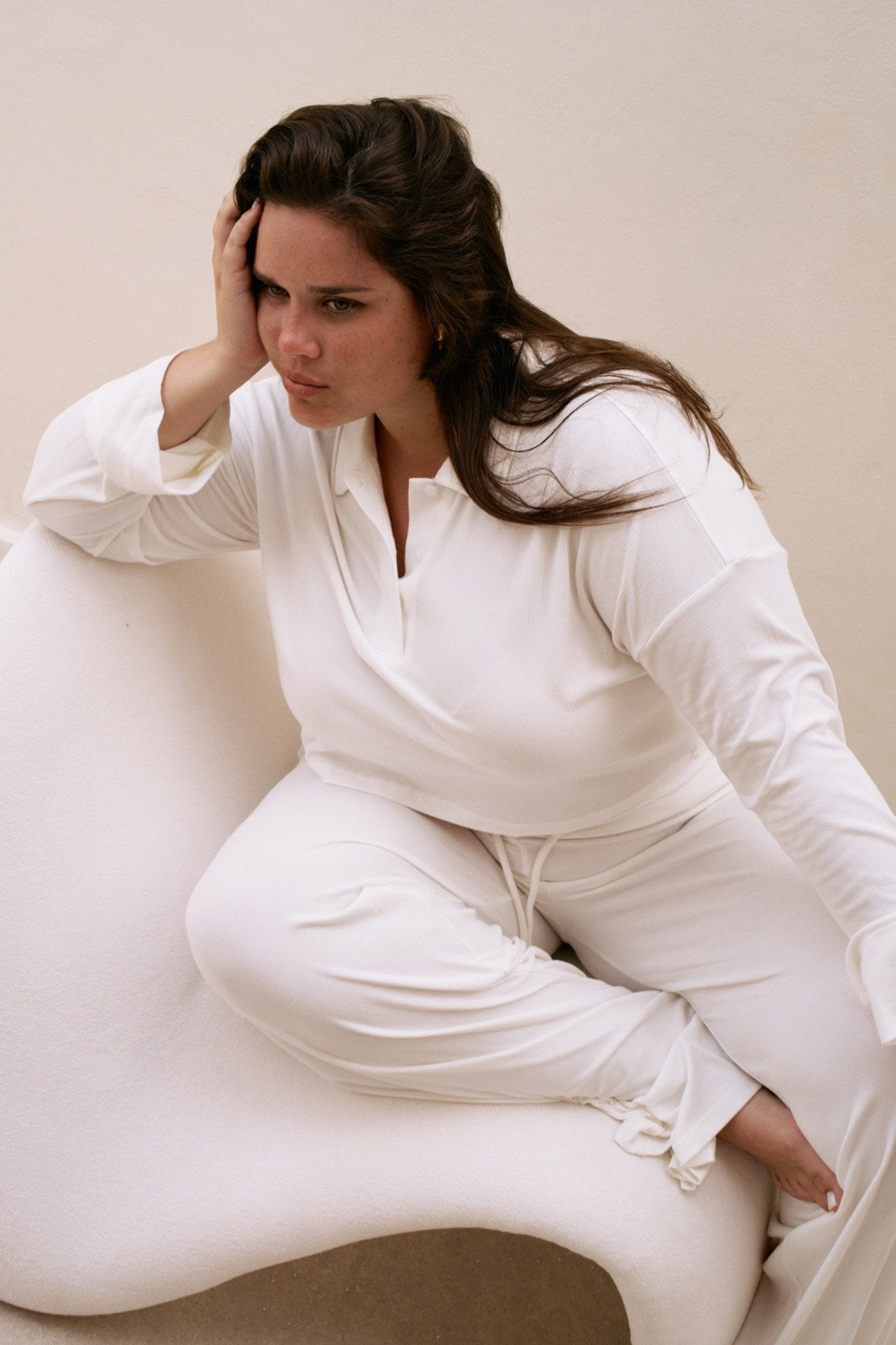 Woman in a white  long sleeve top and pants sitting on a white chair against a beige background