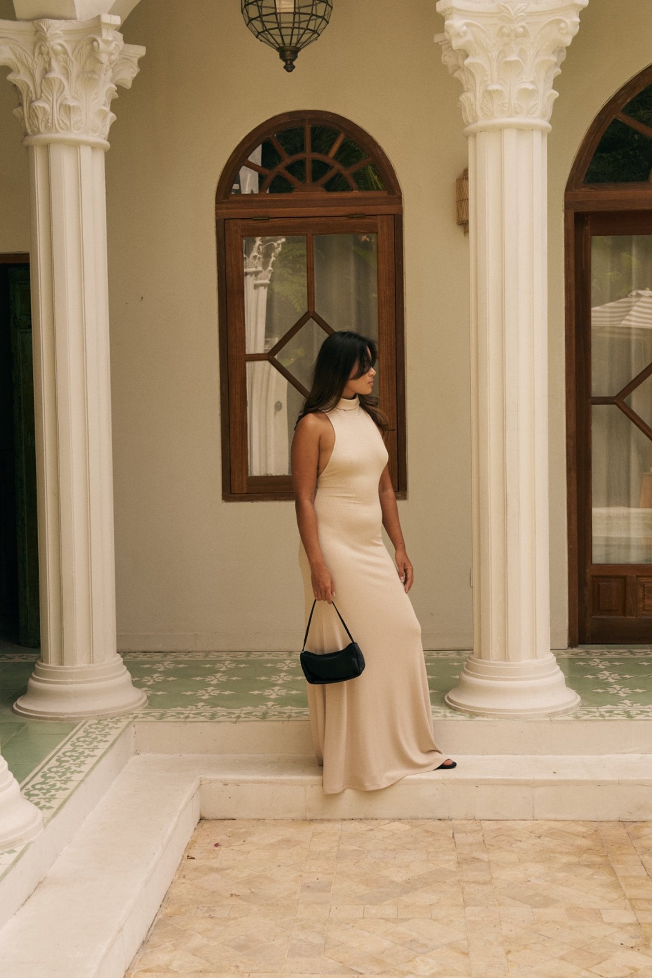 Woman in a beige/coconut dress standing in an elegant interior setting with columns and a mirror.