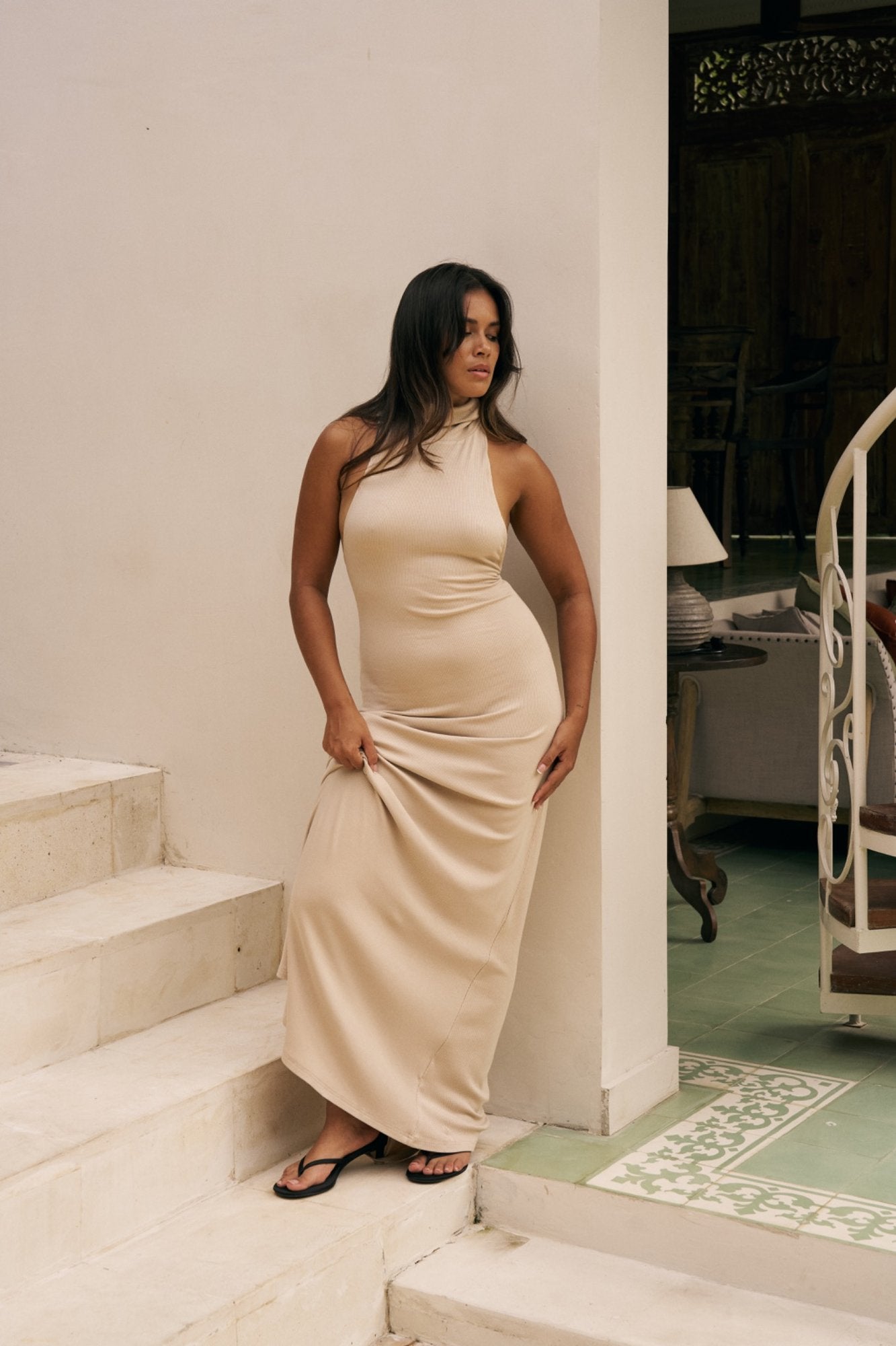 Woman in a beige turtleneck dress standing on a staircase indoors.