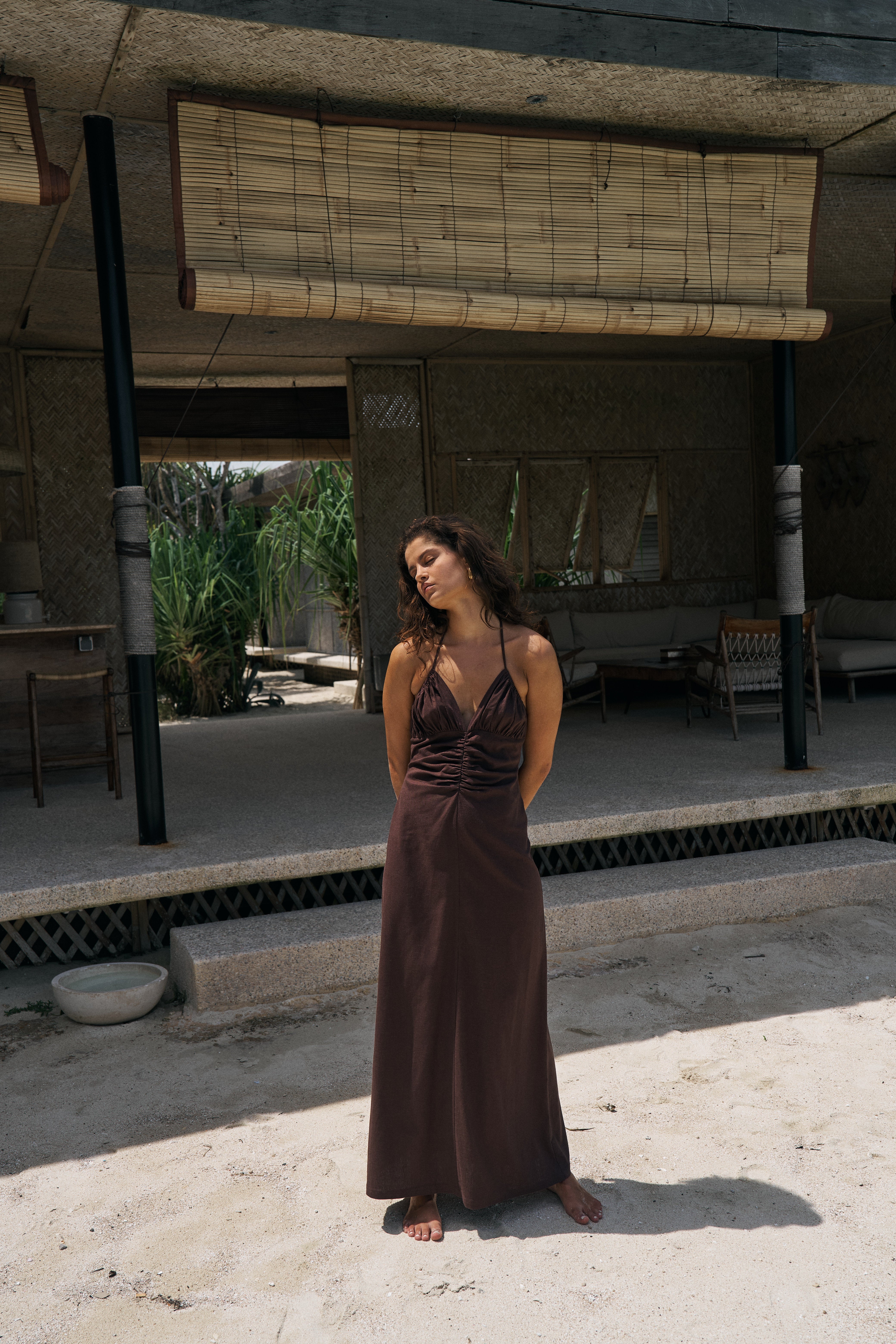 Woman in a brown dress standing in an outdoor setting with a building and plants in the background.