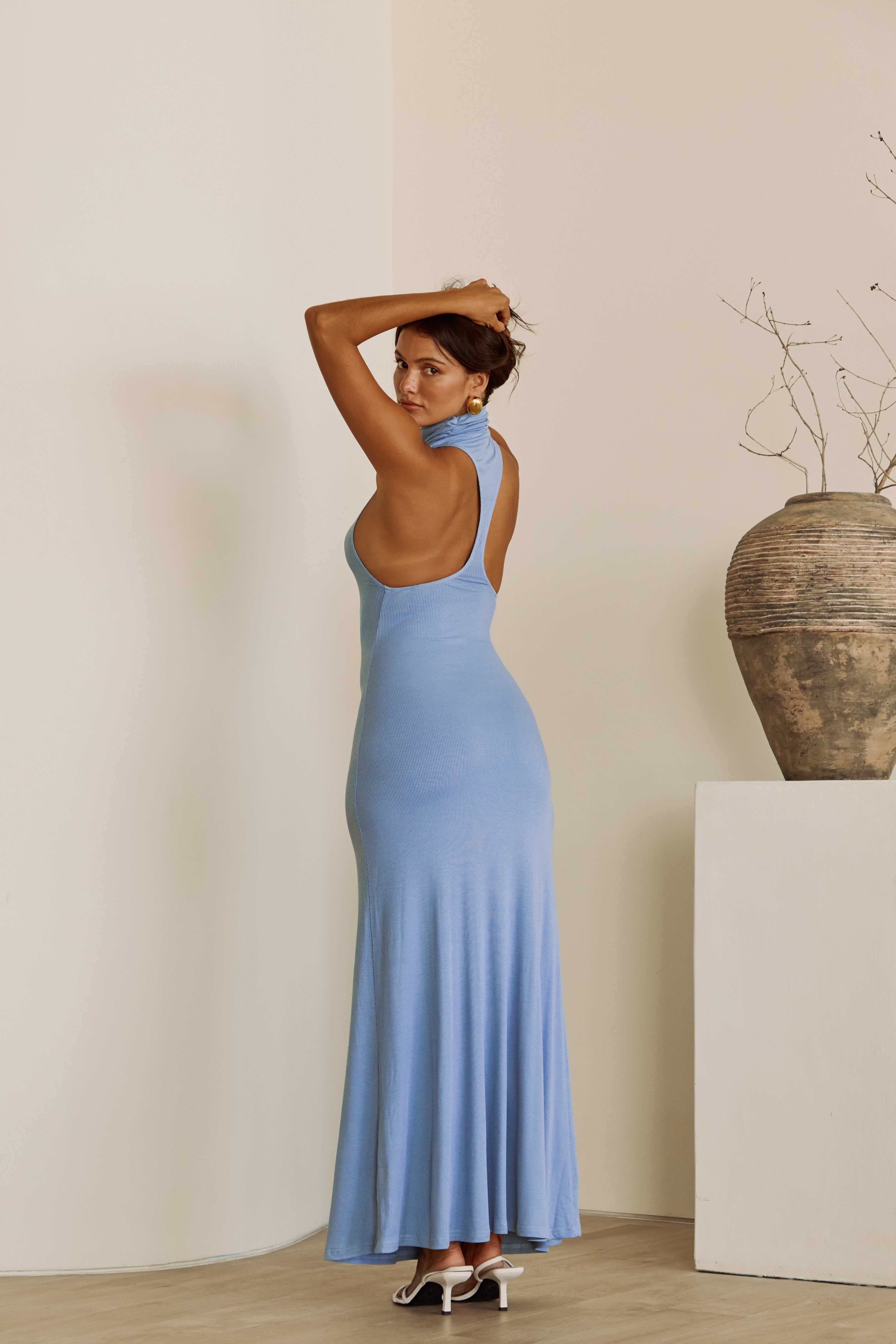 Woman in a blue dress  with turtleneck standing in a minimalistic room with a vase on a pedestal.