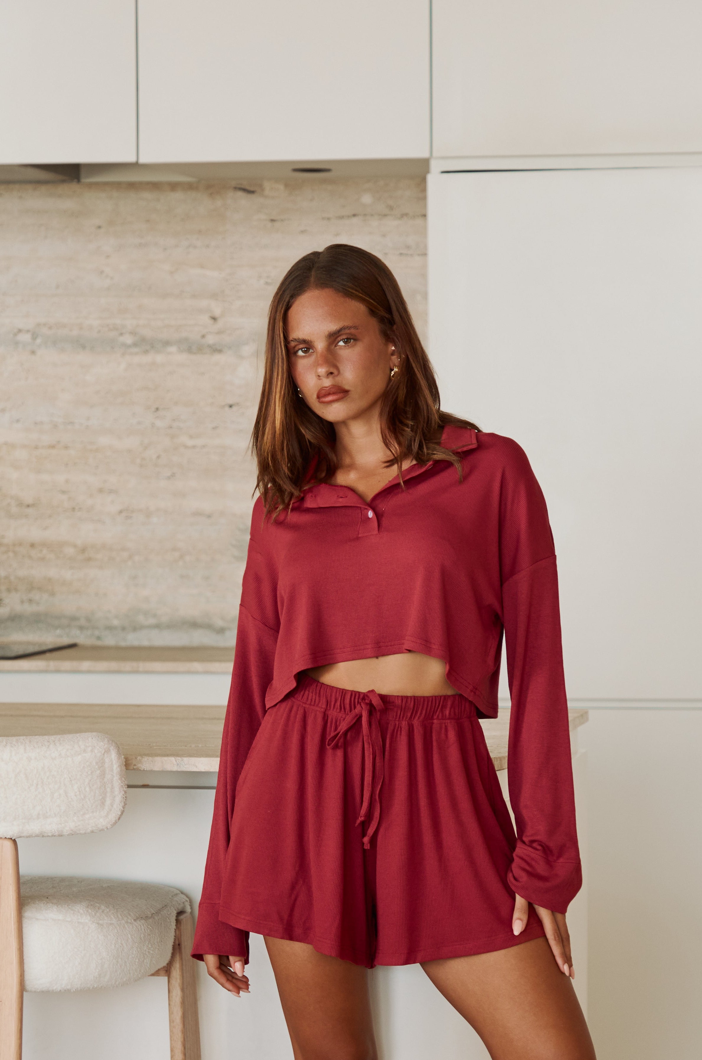 Woman wearing a crimson long sleeve top and shorts standing in a kitchen.