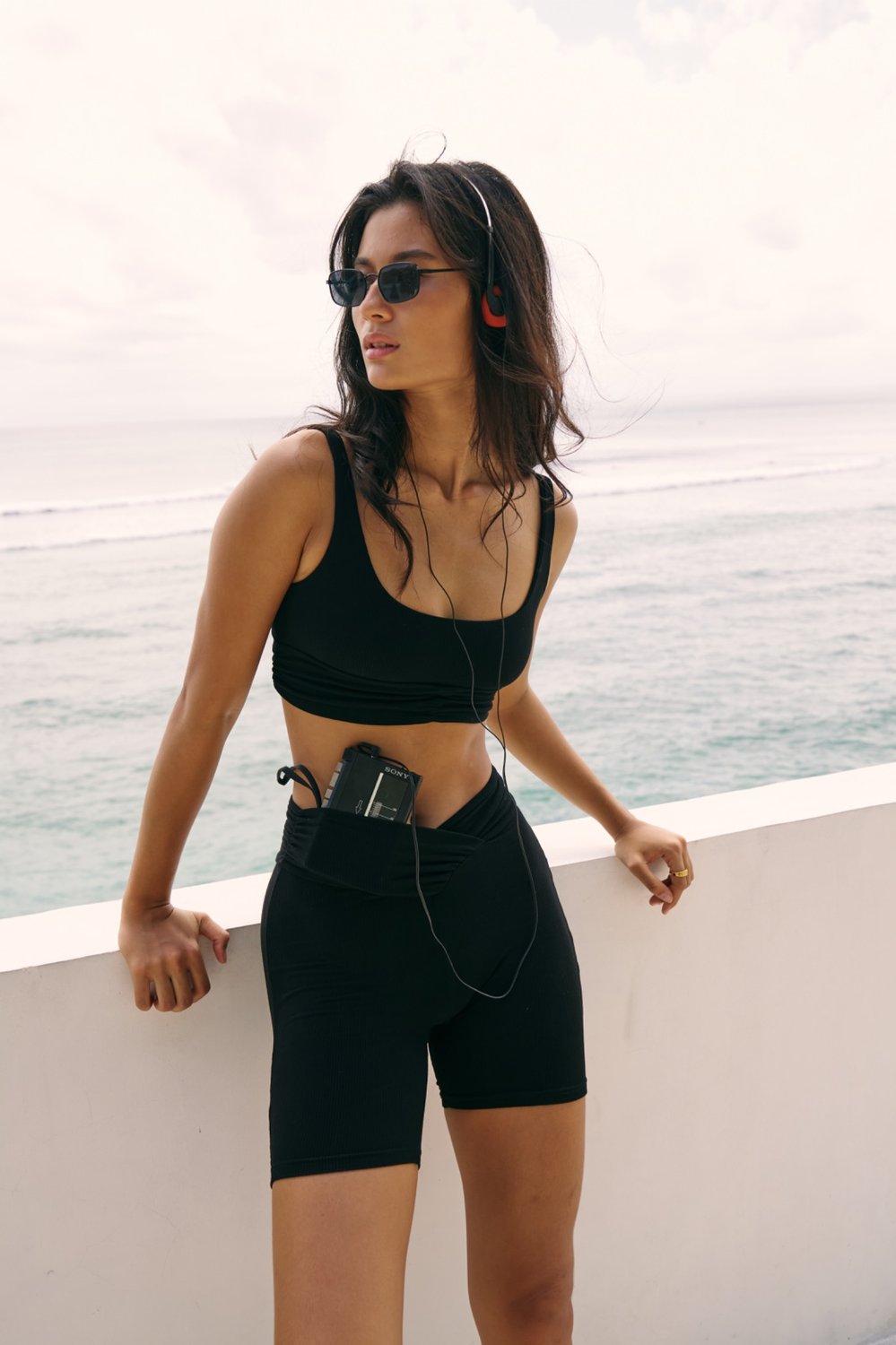 Riot Demi Top and Demi Bottom in black, standing by ocean with walkman accessory in waistband