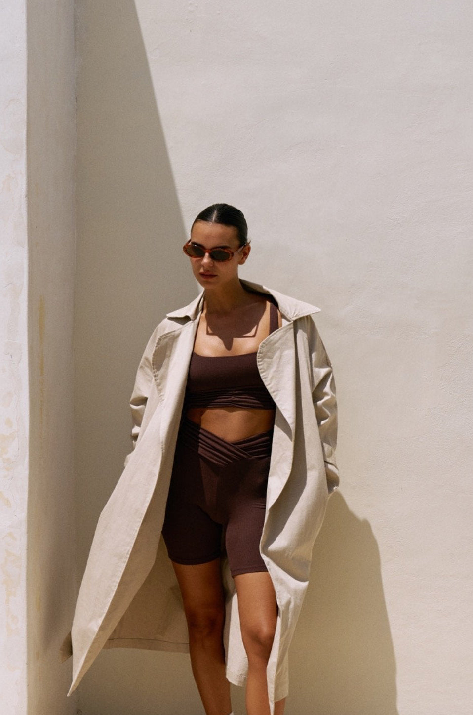 Woman wearing the Demi top and Demi bottom in chocolate, standing against a light textured wall with a long beige trench coat 