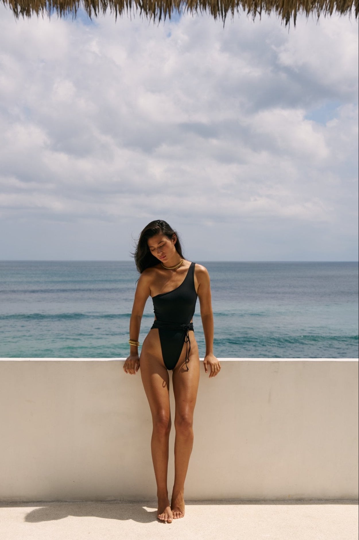 Full-body image of woman standing in the Maddox one-shoulder swimsuit in Black with ocean background.