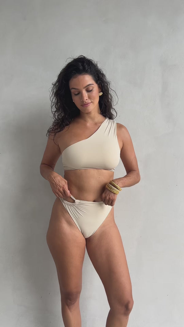 Woman wearing a cream asymmetric bikini top and ruched cutout bottom.