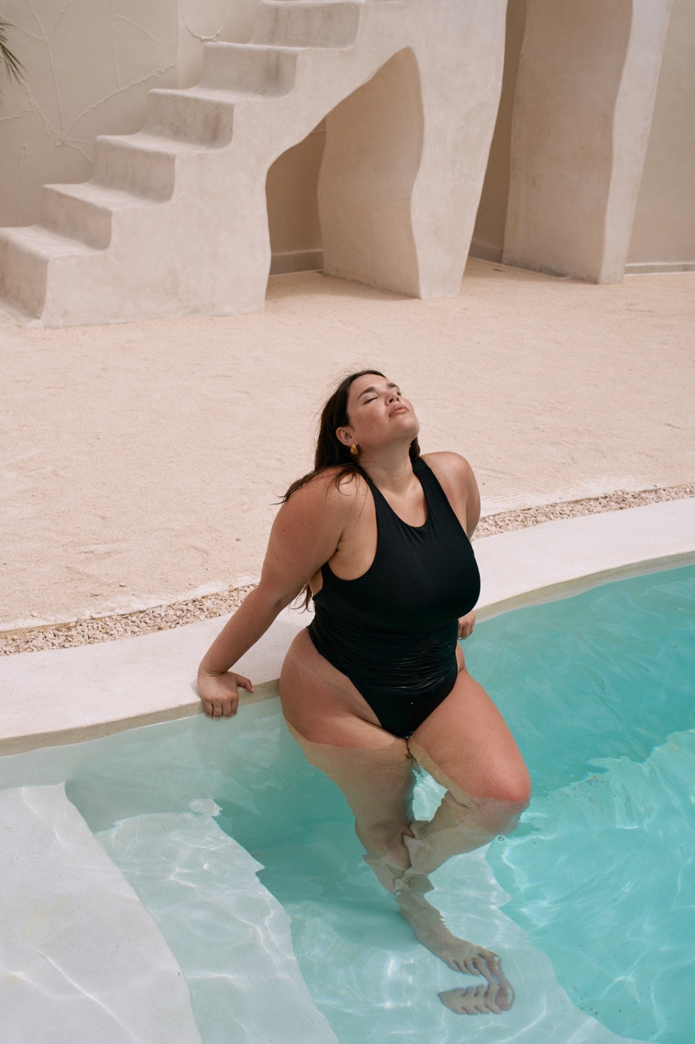A woman wearing a black one-piece swimsuit with ruched detailing lifts herself from a turquoise pool, sunlight highlighting the suit’s texture.