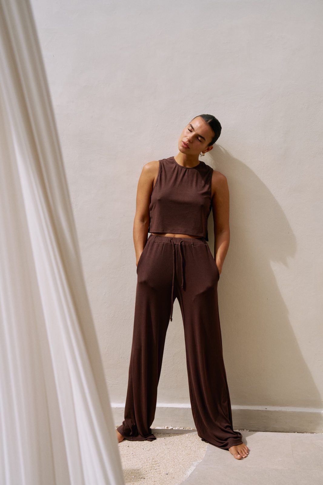 Full body shot of woman standing barefoot in the chocolate brown Simi top and Rory wide leg pants against a neutral wall.