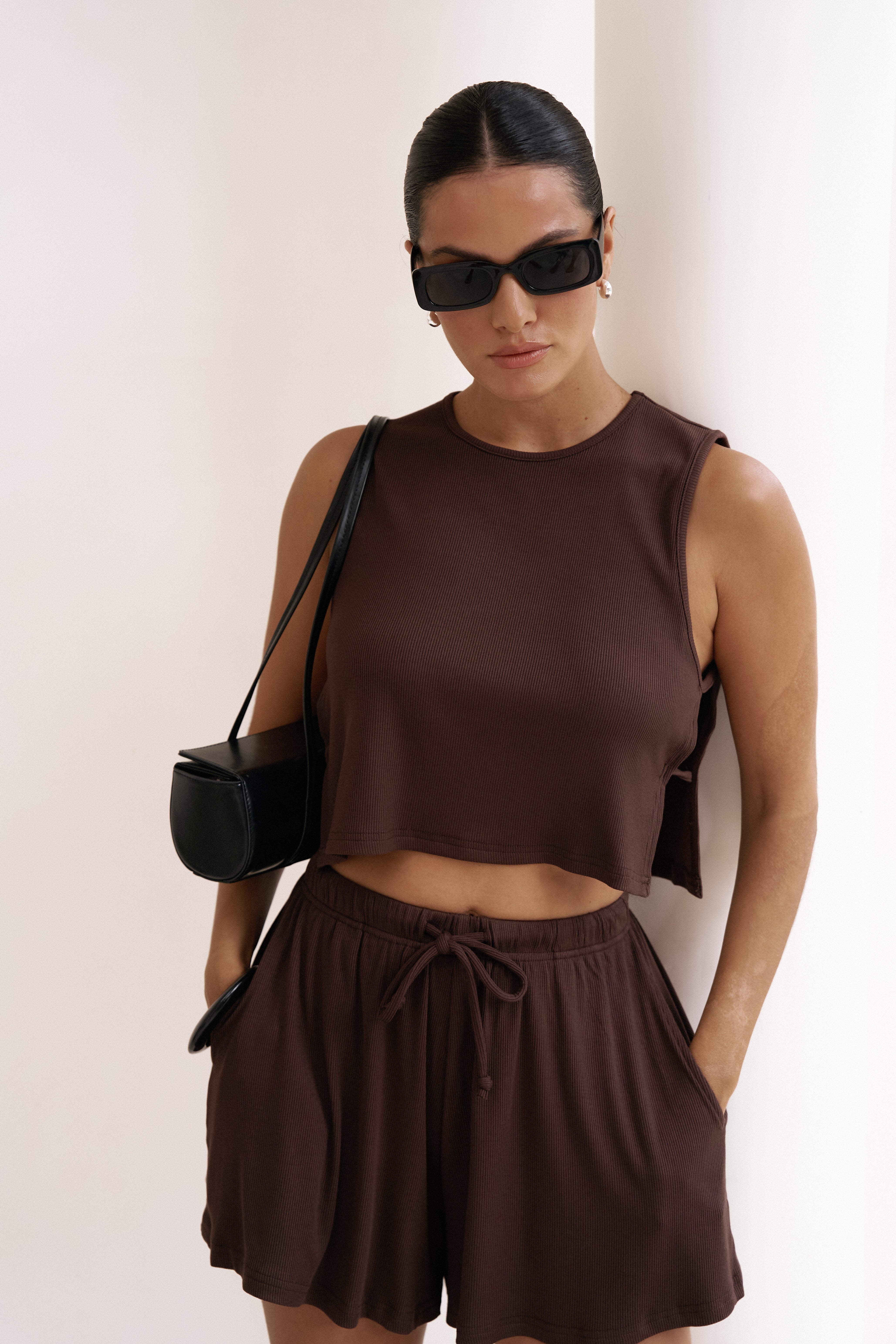Woman wearing a chocolate brown riot swim sleeveless simi top and bottom with a black handbag against a white background